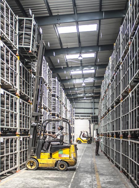 View of a warehouse full of tires stacked high in metal cages and a man in a yellow forklift removing a stack of tires from the very top of the shelves